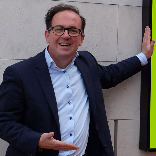 Coaching Aschaffenburg auf dem Farbfoto ist Jürgen Junker Diplom Psychologe Aschaffenburg zu sehen. Er träft einen dunkelblauen Anzug mit hellblauen Hemd und zeigt eine dynamische Geste mit offener Hand. Sein Blick ist im Coaching in Aschaffenburg dem Klienten freundlich zugewandt